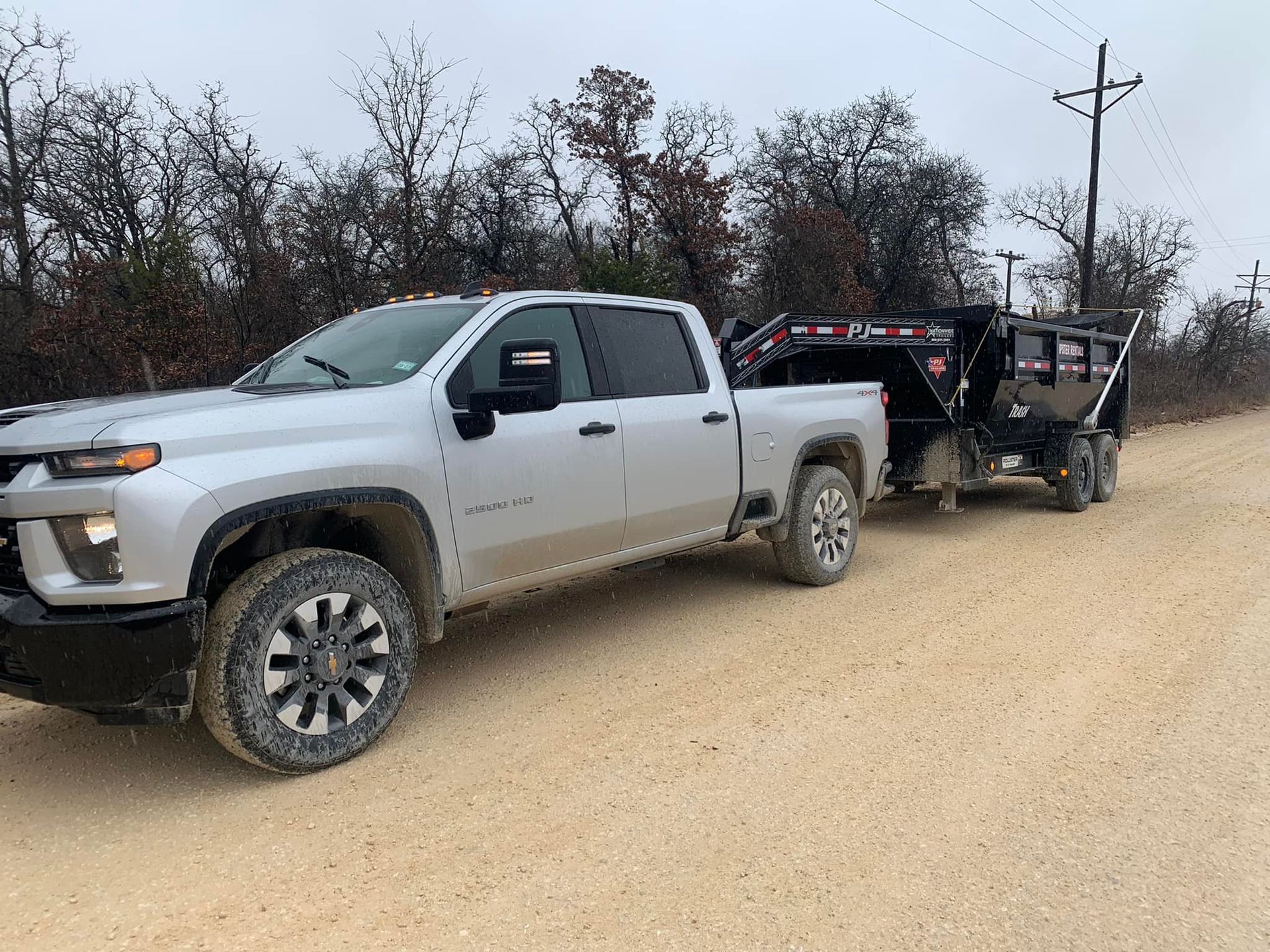 A silver truck is towing a dumpster on a dirt road.