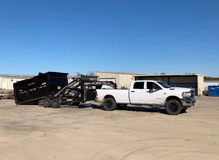A white truck is towing a dumpster in a parking lot.