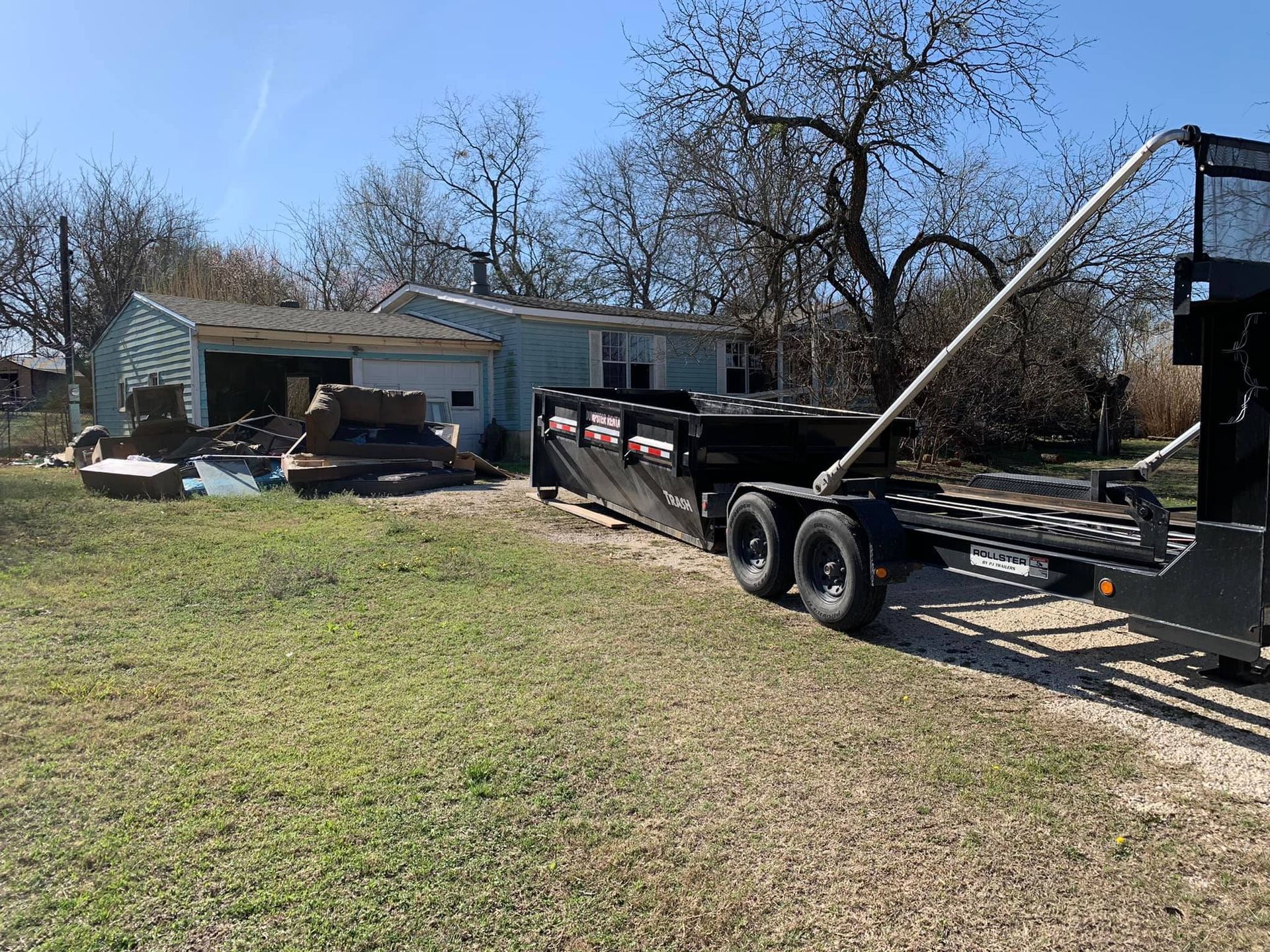 A dump trailer is parked in a grassy field in front of a house.