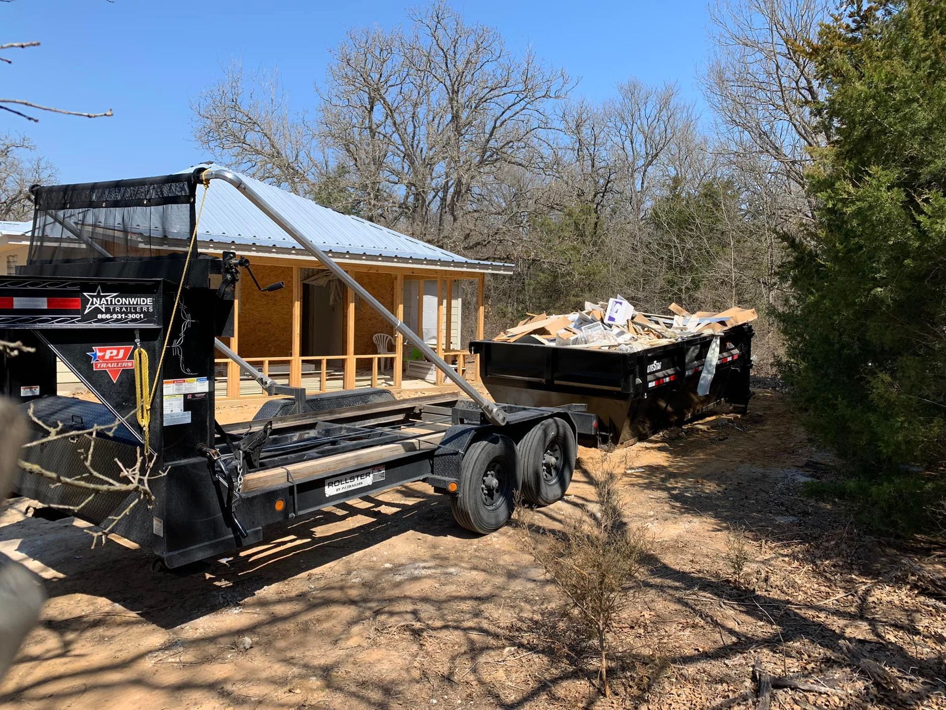 A dumpster is sitting next to a trailer with a house in the background.
