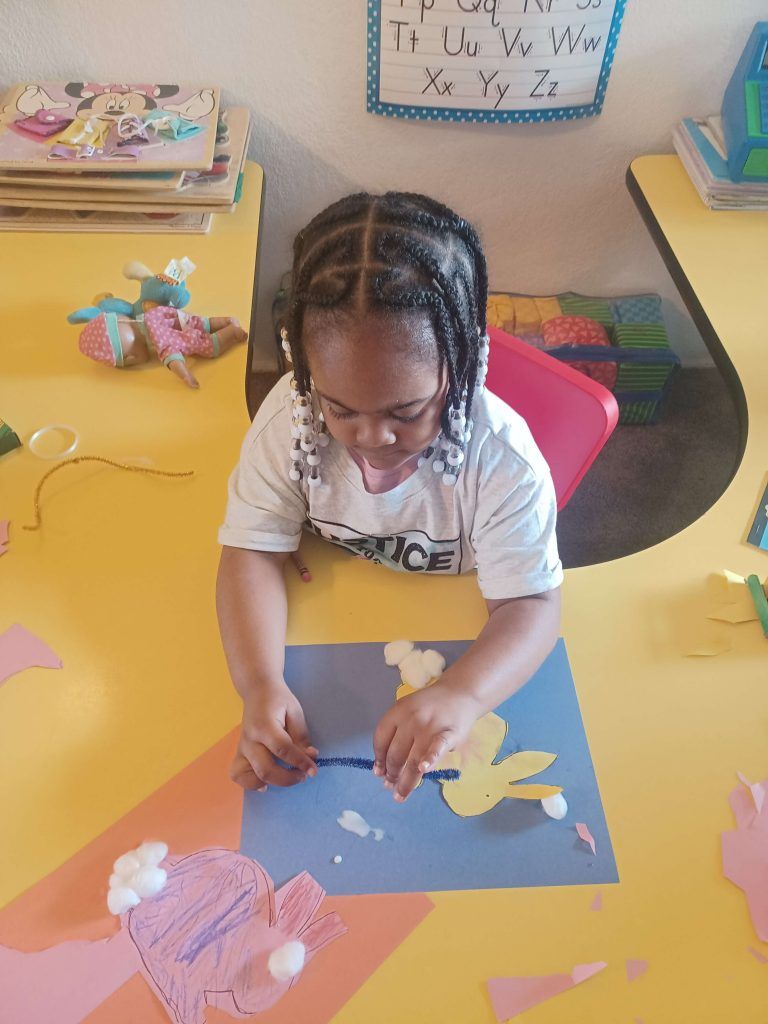 Young person with braids crafting at a yellow table; blue paper, art supplies, and colorful cutouts.