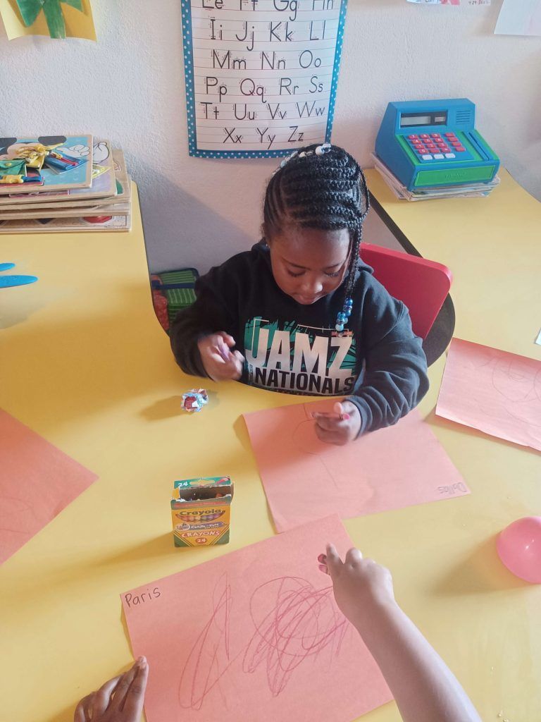 Child drawing at a table in a classroom, with alphabet chart and a toy cash register in the background.