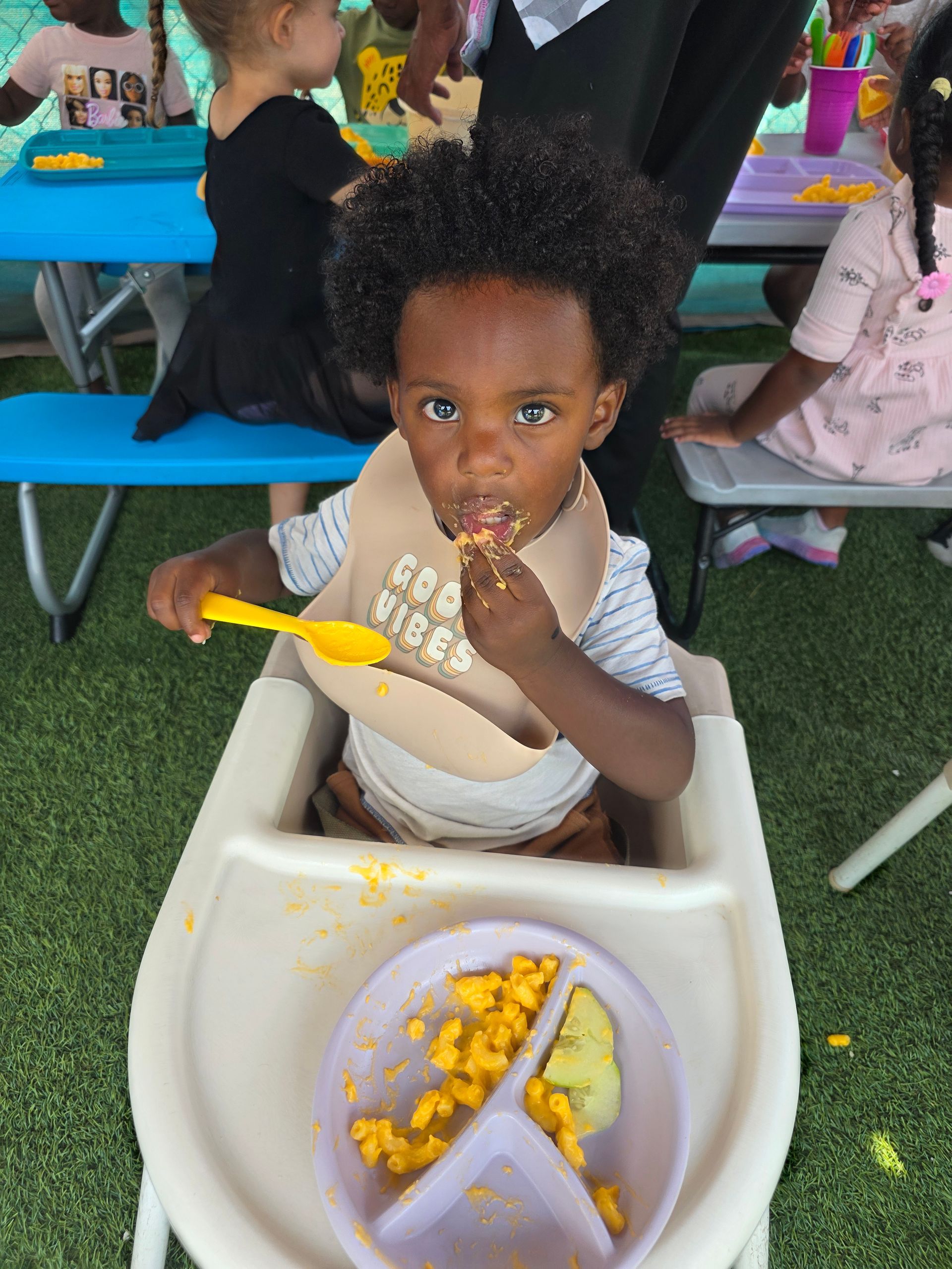 Child in high chair eating mac and cheese, wearing a bib, outdoor setting.