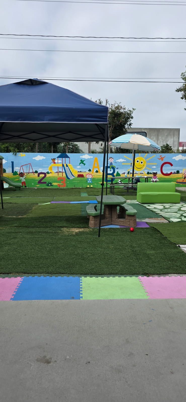 A playground with a painted mural, artificial grass, picnic table, and colorful mats under a cloudy sky.