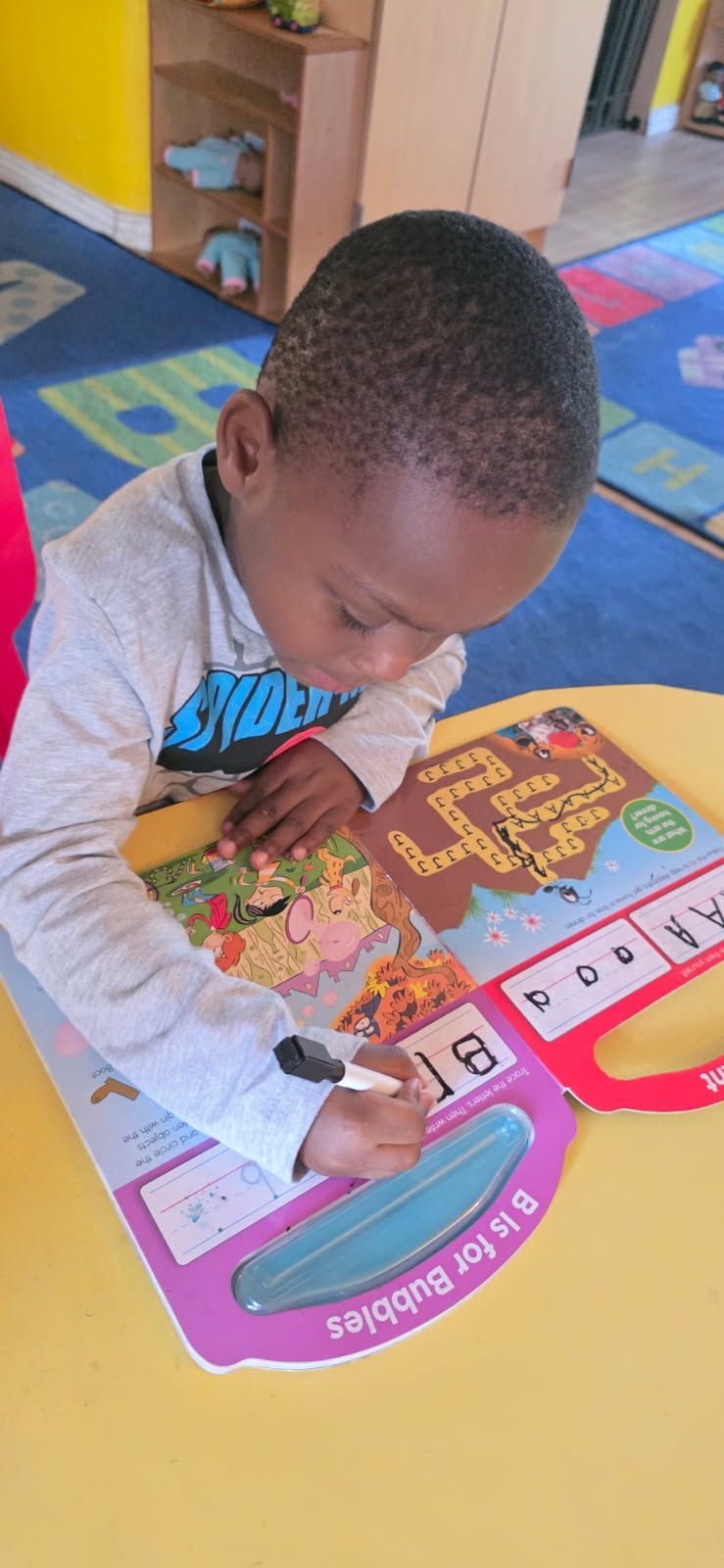 A child writes in a book. The child sits at a yellow table in a classroom setting.
