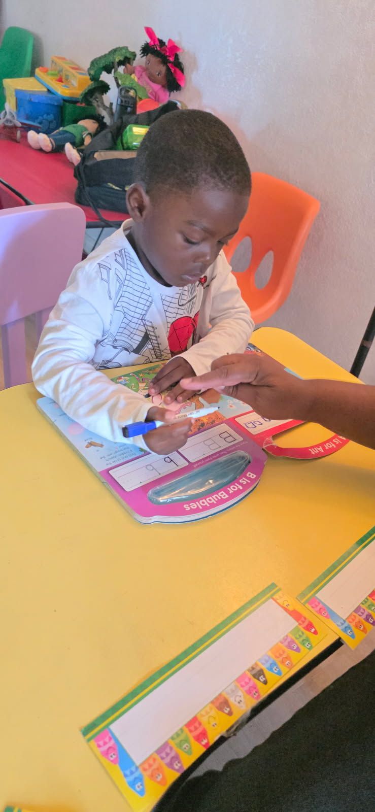 A young child at a table is working on a worksheet with help from an adult's hand.