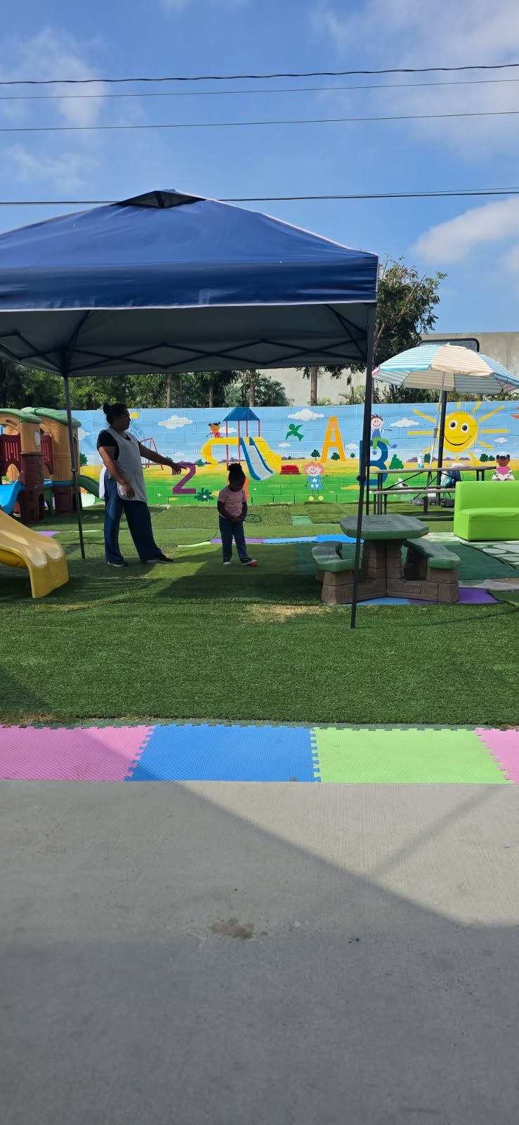 Child playing on playground under a blue canopy. A person watches the child. Colorful painted mural on the wall.