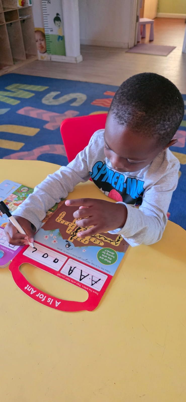 A child sits at a yellow table, holding a wooden stick, playing with an ABC learning toy.