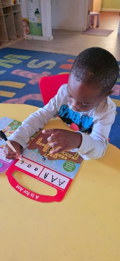 A child sits at a yellow table, holding a wooden stick, playing with an ABC learning toy.