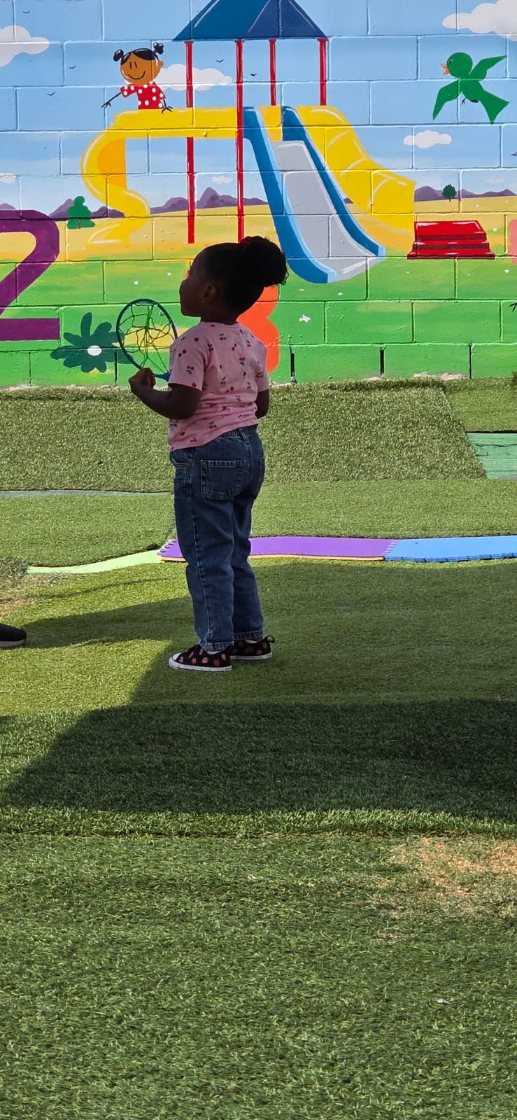 Child standing on artificial turf, holding a badminton racket, with a playground mural in the background.