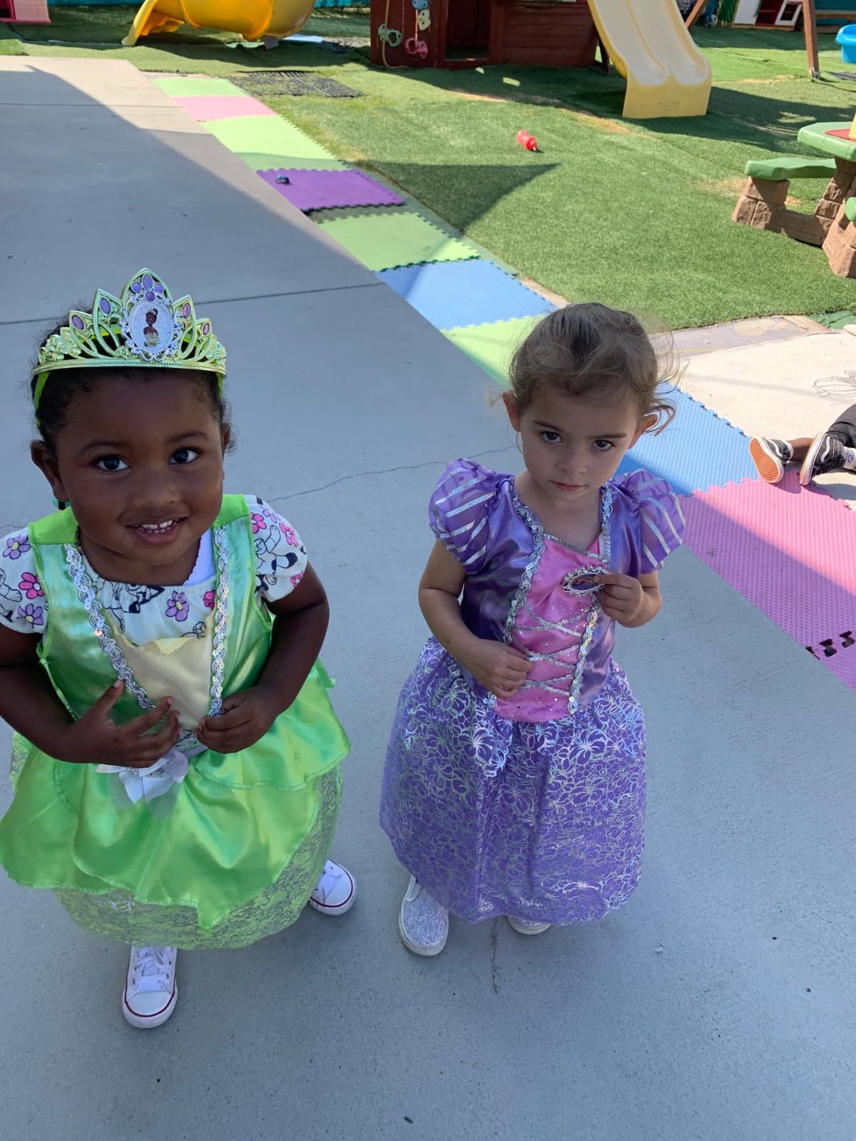 Two young children in princess costumes outside. One in green, the other in purple.