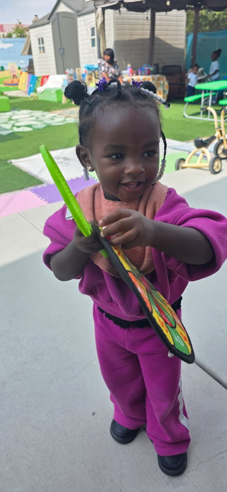 A smiling child in a pink sweatsuit holding a green and black toy outside.