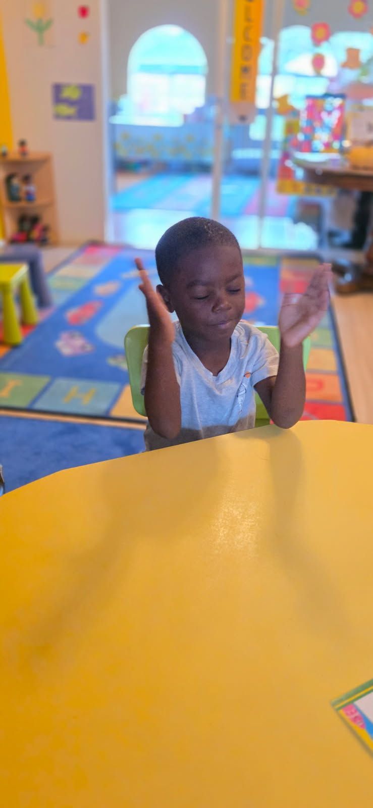 Young child with hands raised sits at a yellow table in a classroom.