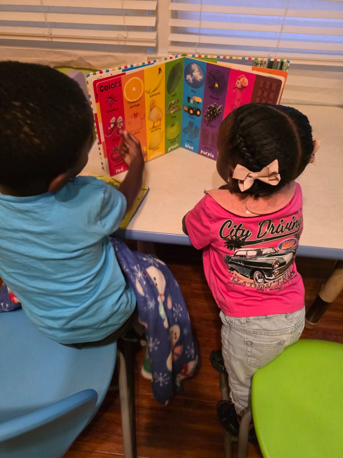 Two children looking at a colorful book on a table. One child is wearing a blue shirt and the other a pink shirt.