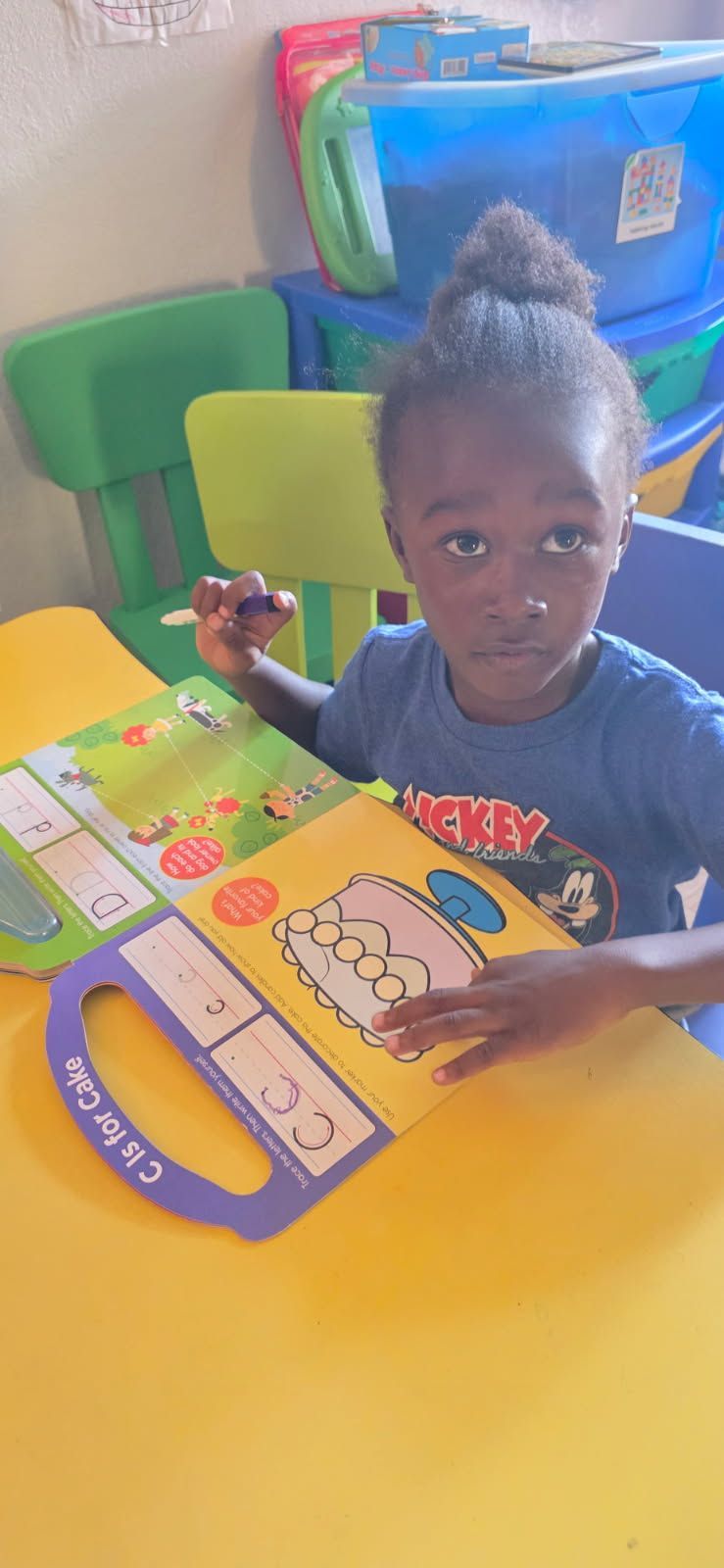 Child seated at a yellow table, holding a pen and looking at a colorful learning book, with toys in background.