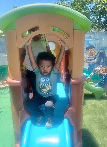 Child on a blue slide, holding onto the top of the play structure. Other children and caregiver in the background.