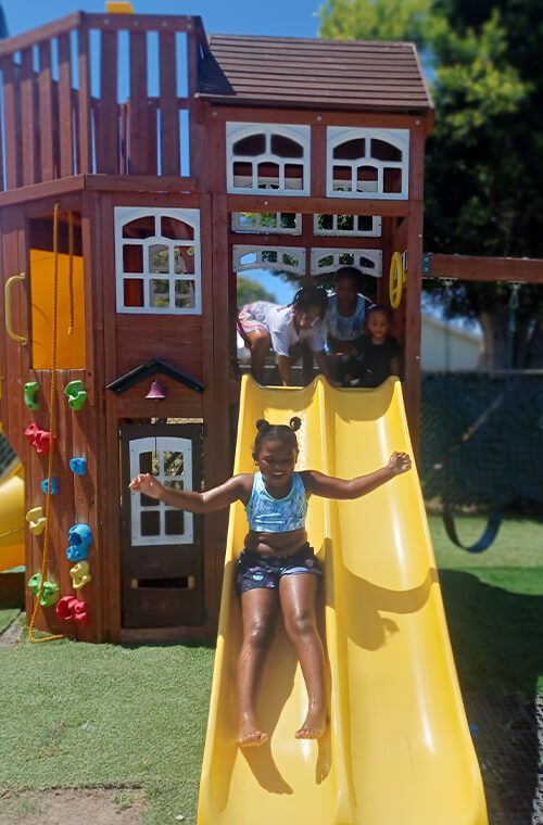 Children playing on a wooden playset with a yellow slide. One slides down, arms outstretched, while others watch from above.