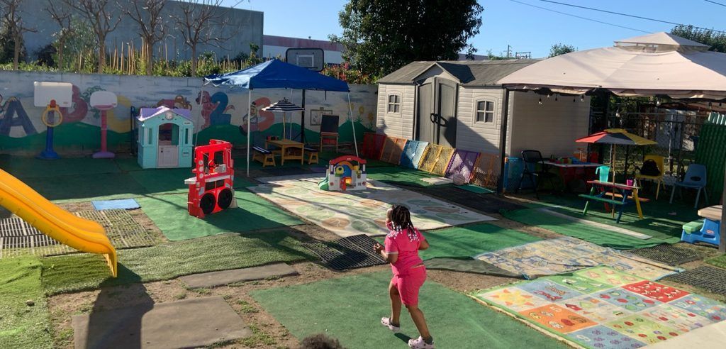 A child in a pink dress runs through a playground with playhouses, a slide, and a shaded seating area.