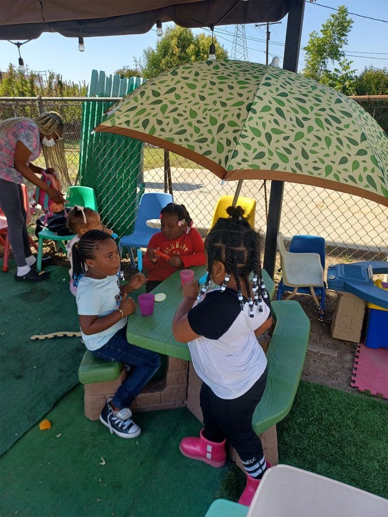 Children sitting at a picnic table under a patterned umbrella, playing outdoors with an adult nearby.
