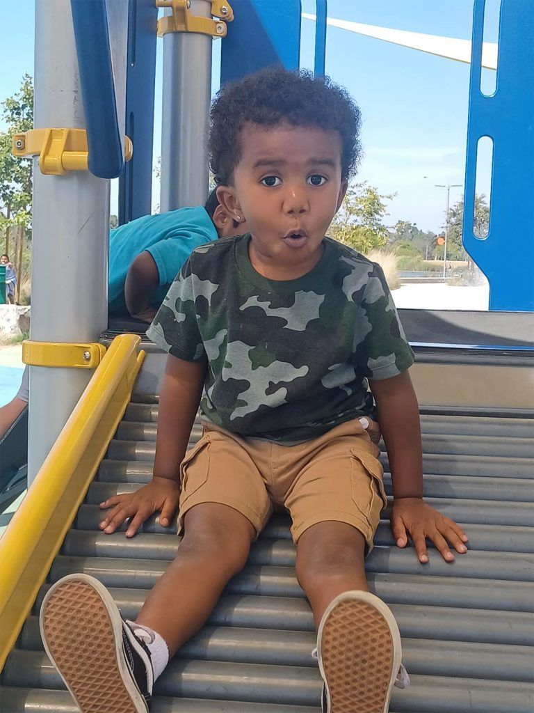 Young child with curly hair on a playground slide, making a face. Wearing a camo shirt and khaki shorts.