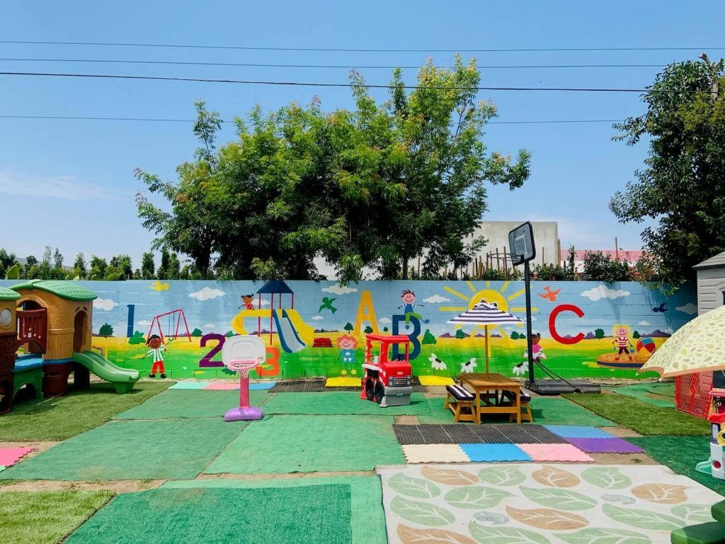 Playground with painted wall, play structures, and a basketball hoop on a green surface under a blue sky.
