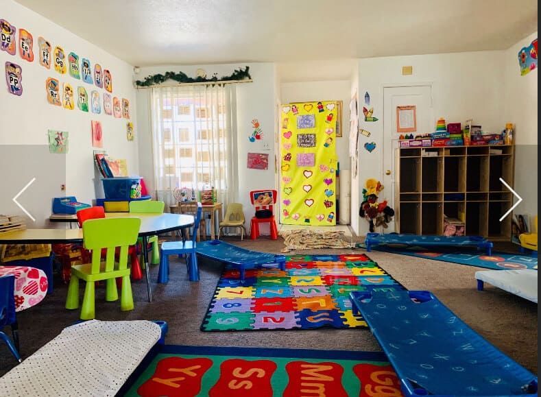 A brightly decorated preschool classroom with tables, chairs, cubbies, mats, and cots.