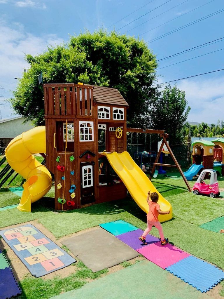 Child running toward a wooden playset with a yellow slide, swing set, and hopscotch tiles on a sunny day.