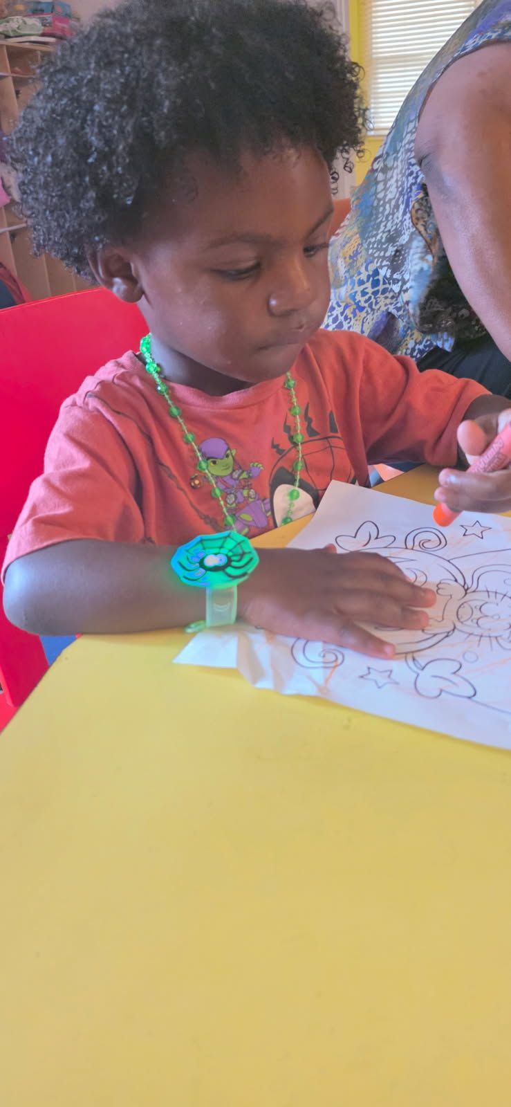 A child with curly hair coloring at a yellow table; they wear a necklace and a flower-shaped watch.