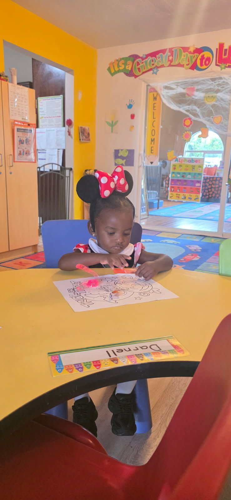 A child with Minnie Mouse ears draws at a yellow table in a classroom, with colorful decorations.