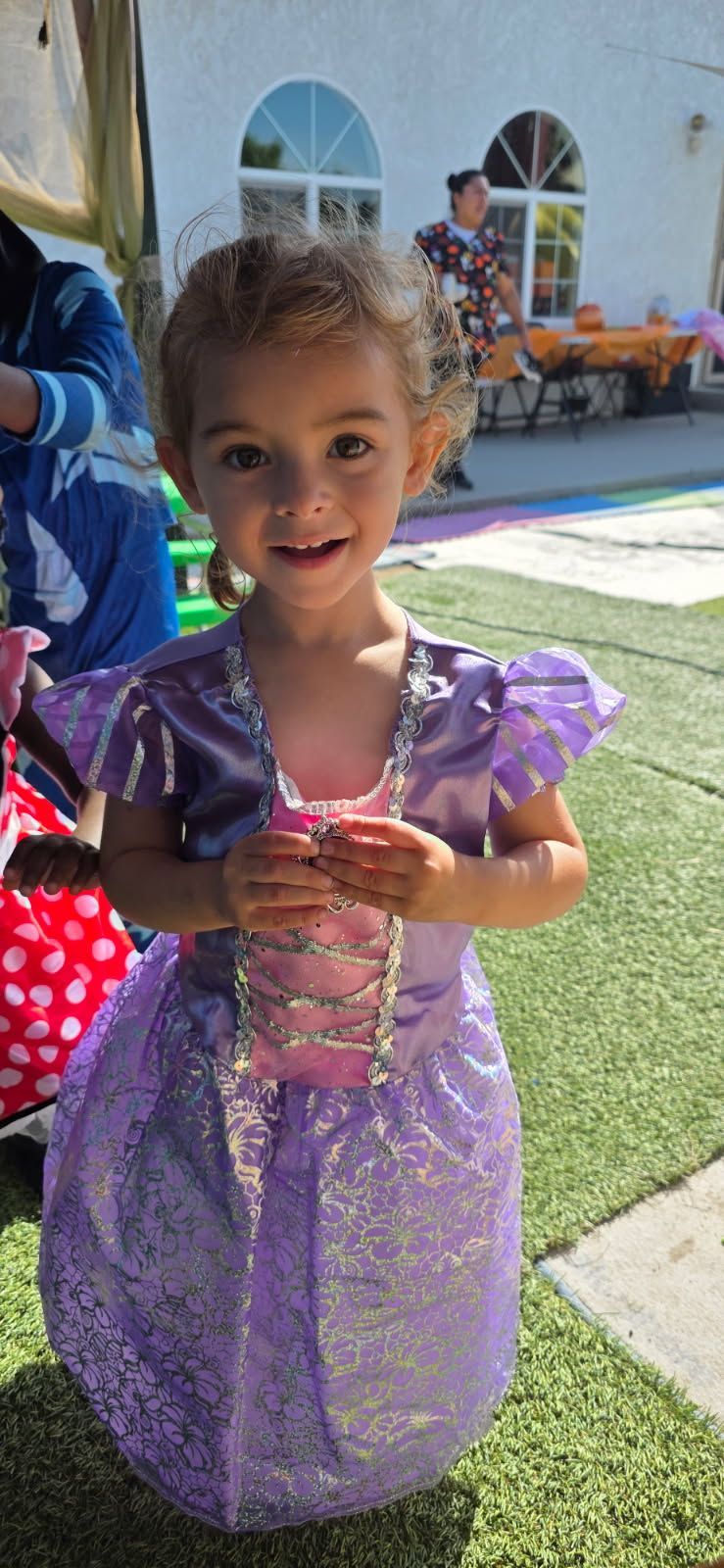 Young child in a purple princess costume smiles at the camera outdoors, with a white building and grass visible.