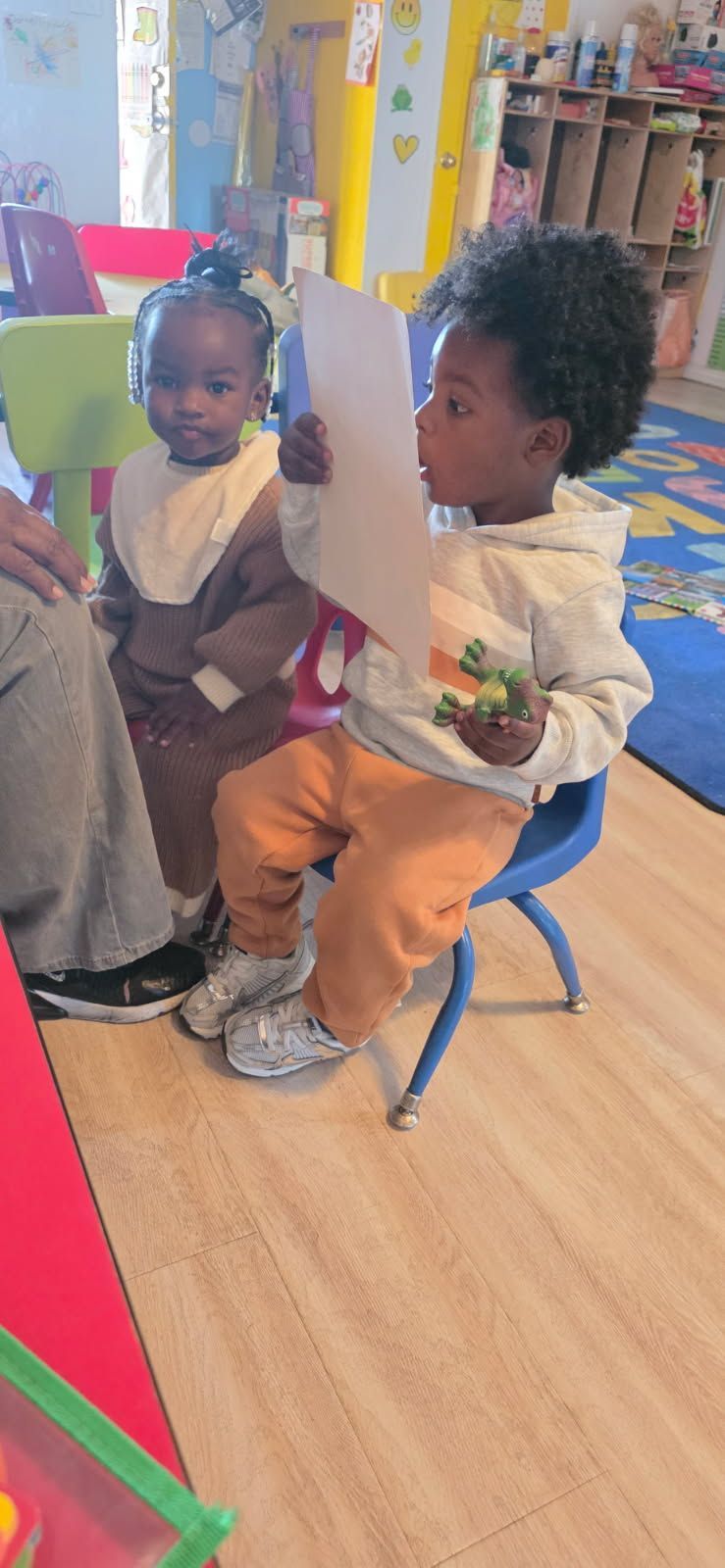 Two young children in a classroom. One child reads a paper, the other watches.