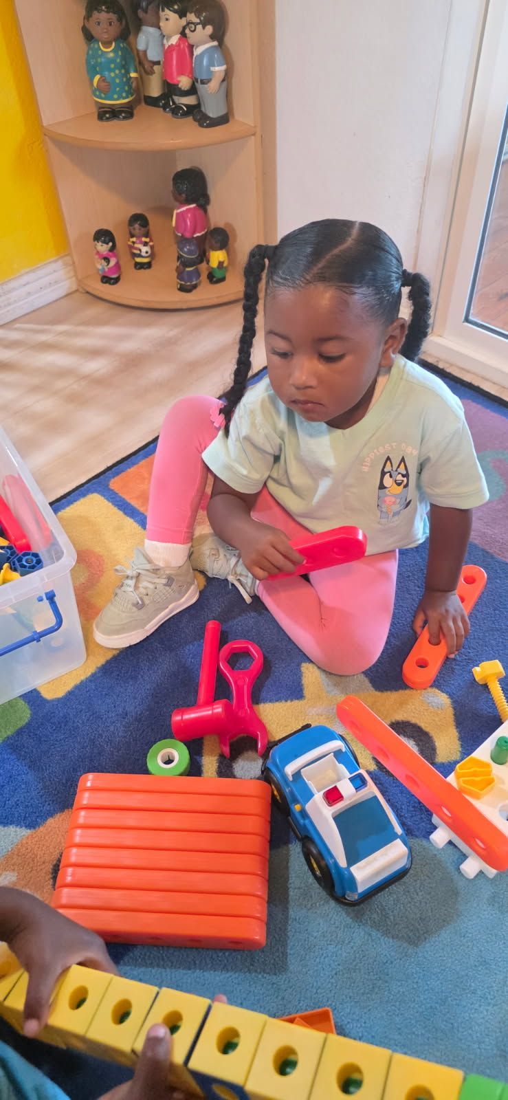 A child sits on a blue rug playing with toys. She has pigtails, wearing pink pants and a light blue shirt.