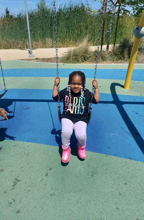 Girl swinging on a playground swing, smiling. Blue and green ground, sunny day.