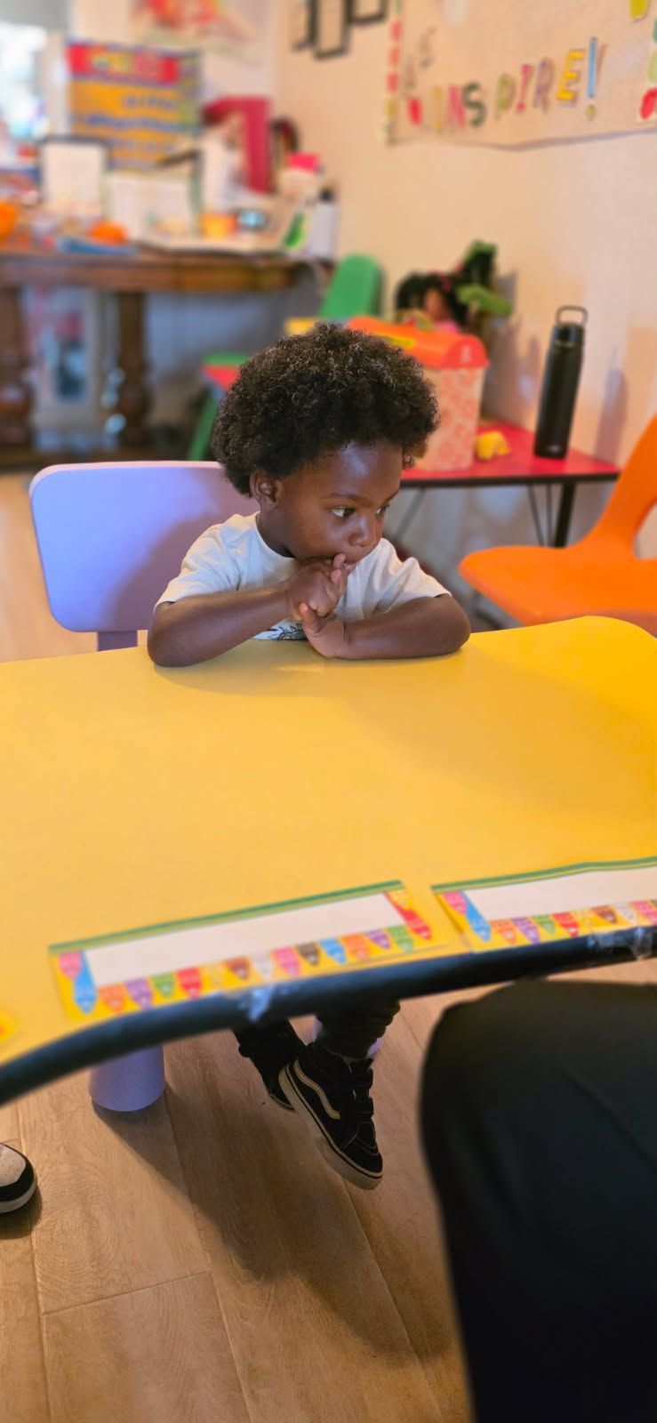 A child seated at a yellow table with hands clasped. They are wearing a white shirt and looking down. The setting appears to be a classroom.