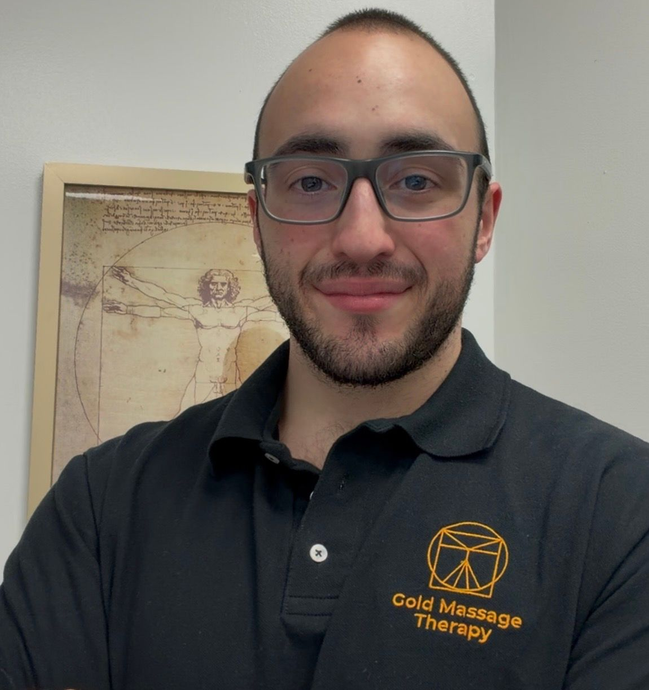 Man wearing glasses and a black polo with a gold logo, smiling indoors beside framed wall art