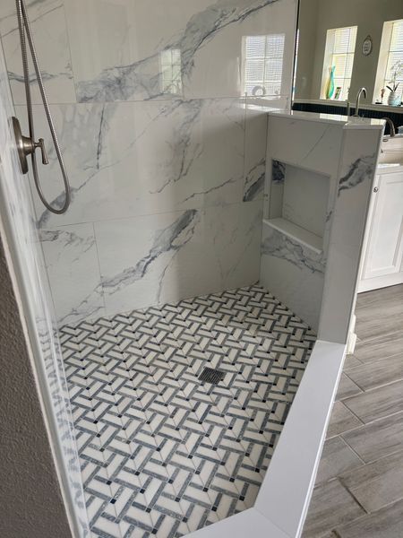 A corner shower with white and blue marble-look wall tiles, a mosaic herringbone floor, and a built-in shelf.