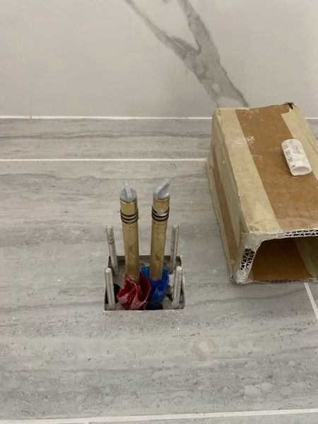 Pipes protruding from a tiled floor with red and blue tape, set in a metal enclosure. A cardboard box rests nearby.