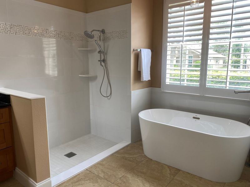 Bathroom with walk-in shower, freestanding tub, and window with shutters. Light tan walls and beige tile floor.