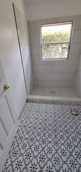 Bathroom with white and gray patterned tile floor, tiled shower, and a window.