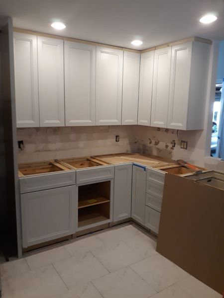 White kitchen cabinets installed in a corner, with unfinished countertops and drywall.