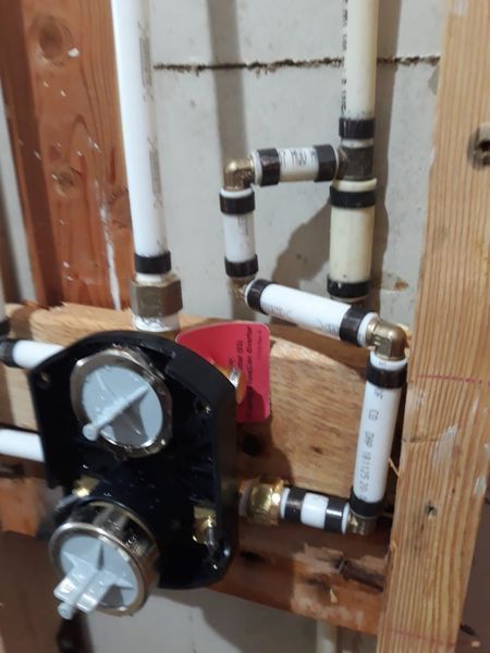 Plumbing pipes and a shower valve installed within a wall frame, featuring white pipes, black fittings, and a black valve.