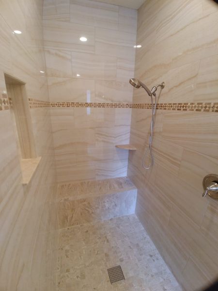 Tiled shower stall with a built-in bench and shelf. Beige and brown tones. Showerhead and drain visible.