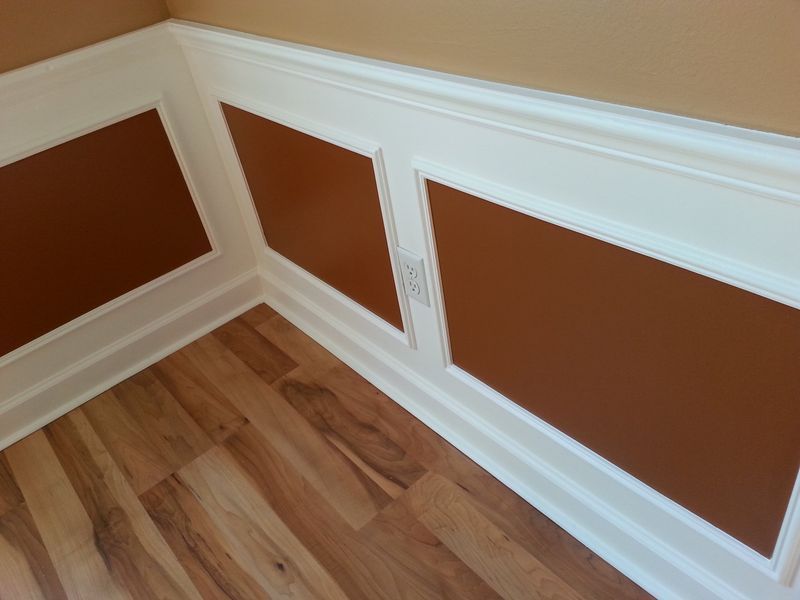 Corner of room with brown panel wainscoting framed in white molding, with a power outlet.  Light wood flooring.