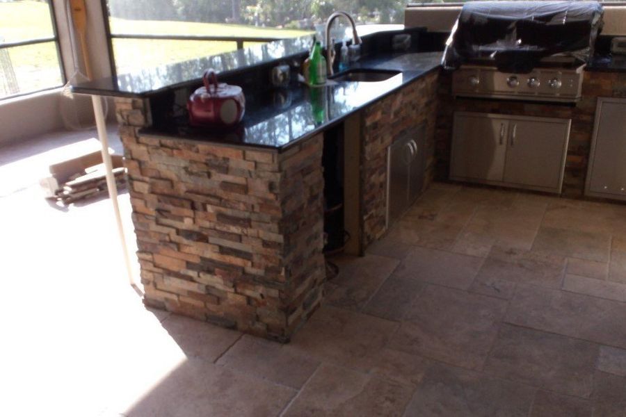 Outdoor kitchen island with stone facade and black countertop. Sink, grill, and storage visible.