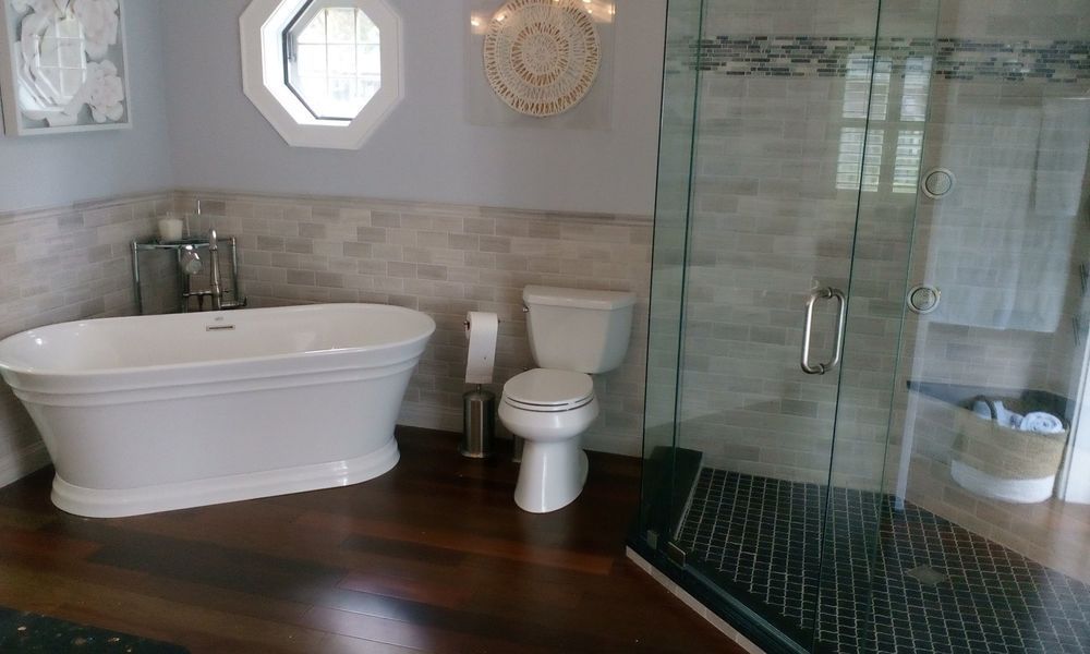 Bathroom with white soaking tub, toilet, and glass shower, with dark wood flooring and light-colored walls.