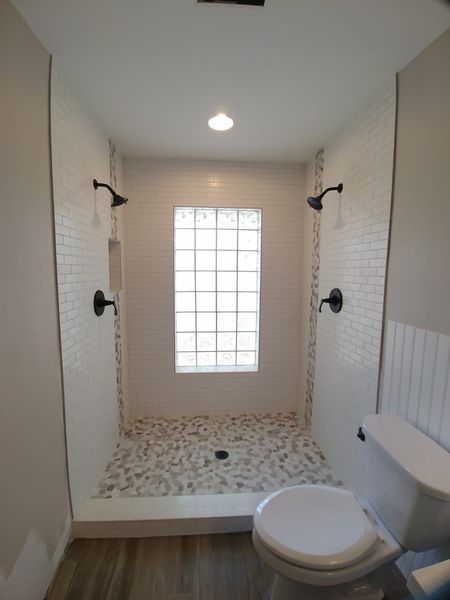 Bathroom with dual showerheads, glass block window, mosaic shower floor, and toilet. White and neutral tones.