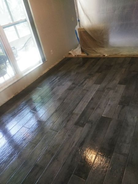 Newly installed wood-look tile floor in a room with a window, reflecting light, with plastic covering nearby.