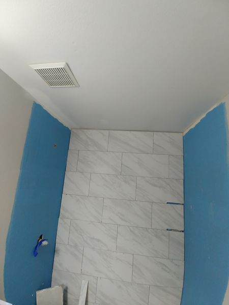 Bathroom under construction with white tiled wall and blue surrounding walls. Ceiling has a vent.