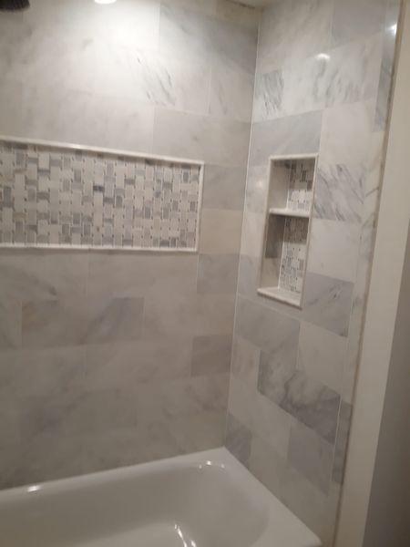 Bathroom shower with white marble-like tile, built-in shelves, and a decorative tile accent. A white tub is visible.