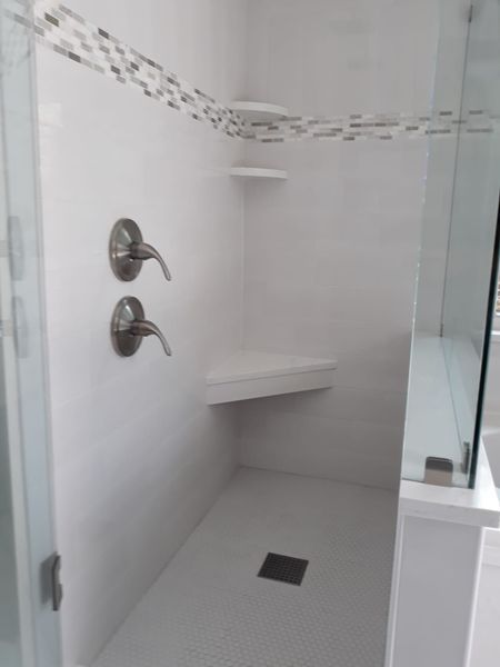 White tiled shower with glass door, fixtures, built-in seat, and shelf.